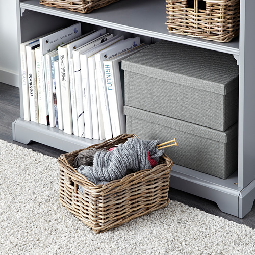 Una cesta de mimbre sobre una alfombra, llena de hilo y agujas de tejer, junto a una estantería gris con libros, cestas tejidas y cajas de almacenamiento en estantes.