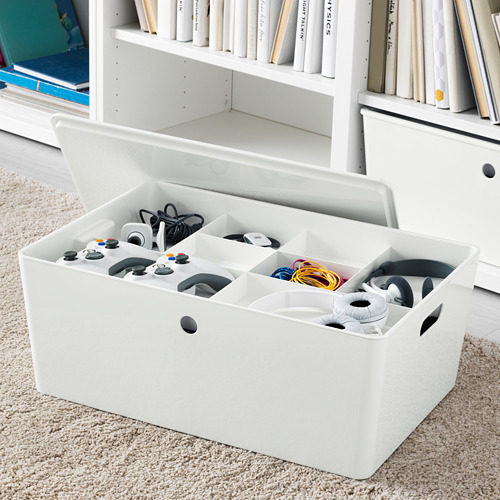 Caja organizadora blanca con múltiples compartimentos que contienen controladores de juegos y cables, colocada sobre una alfombra cerca de la estantería.
