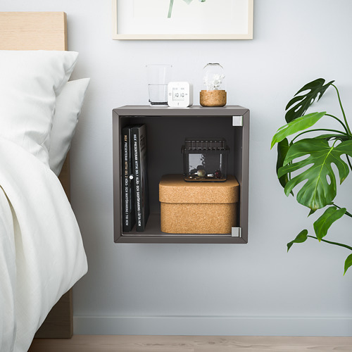 Moderno mueble de almacenamiento cúbico de pared EKET gris con capacidad para libros, cajas de corcho y decoraciones. Cerca de la cama, con una planta verde cerca.