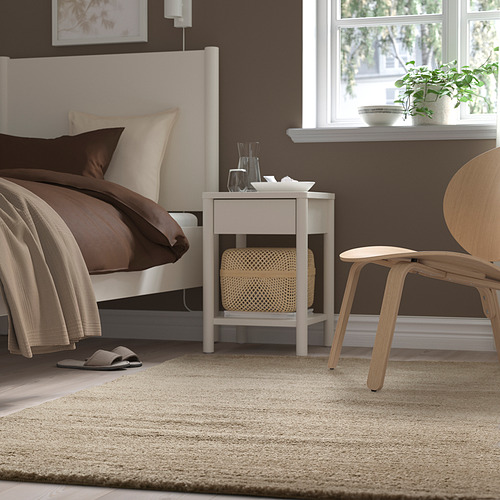 Dormitorio con cama blanca, sábanas marrones, alfombra blanca, mesita de noche blanca y silla de madera.