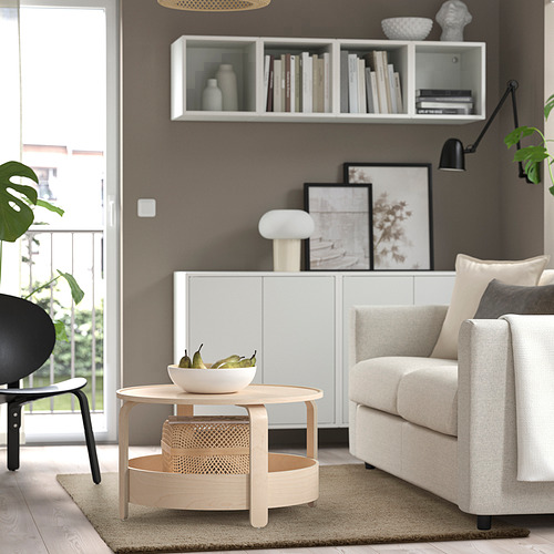 Salón moderno: alfombra neutra, mesa de centro redonda con frutas, sofá beige, silla negra y estantería.