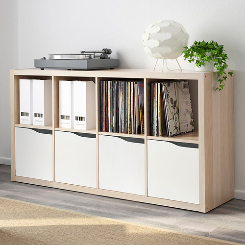 Estantería KALLAX de color claro llena de discos de vinilo y cajas de almacenamiento. En la parte superior se encuentra una plataforma giratoria y se colocan una lámpara blanca y una planta al lado.