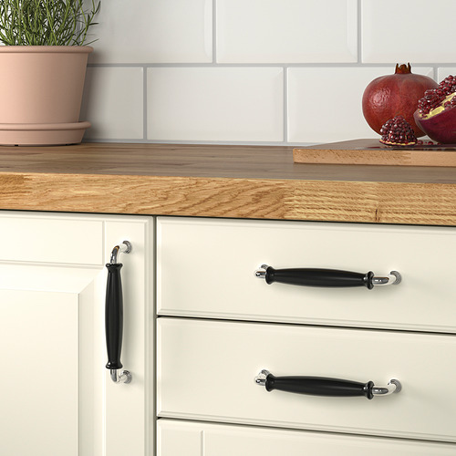 Cajones de cocina con pomos de porcelana negra, armarios blancos, encimera de madera, planta en maceta y granadas frescas.