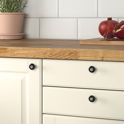 Cocina con cajones blancos con pomos de porcelana negra KLINGSTORP para facilitar el agarre. Encimera de madera con maceta de plantas y granadas.