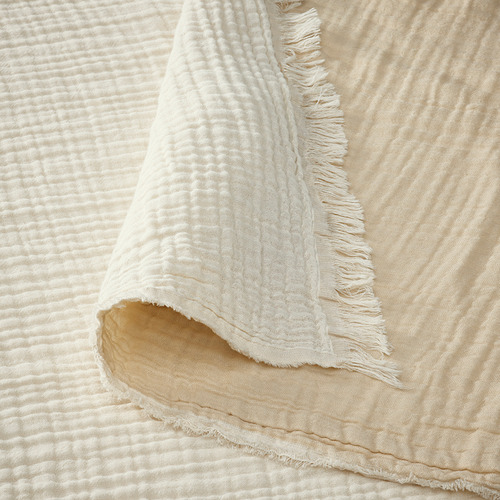 Manta de algodón suave y arrugada con bordes sin rematar, doblada sobre fondo crema.