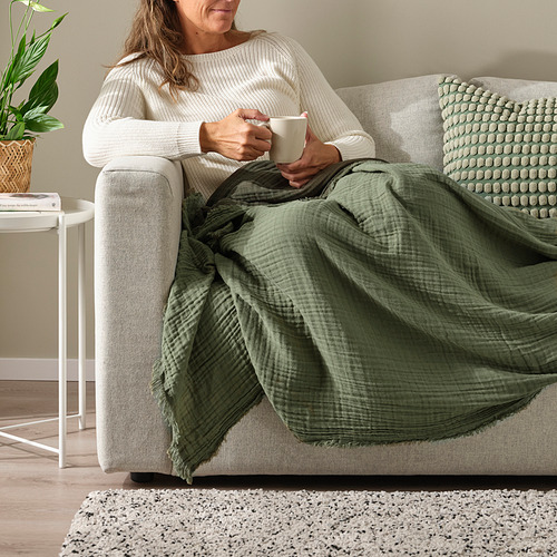 Una mujer se relaja en un sofá beige, sosteniendo una taza de café. Está envuelta en una manta verde de VALLKRASSING.