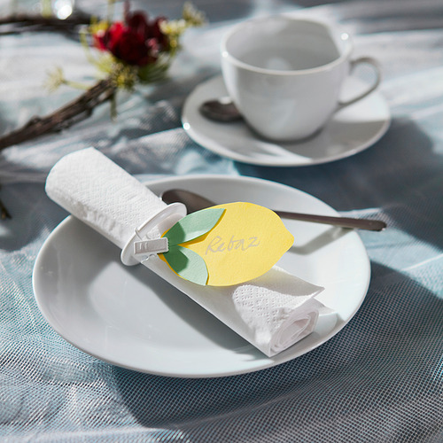 Plato blanco con servilleta enrollada, cartón limón y cuchara; taza y flores en el fondo.
