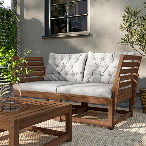 Escena de patio exterior con muebles NÄMMARÖ de acacia marrón, que incluyen un sofá y una mesa de centro con cojines blancos, colocados sobre una alfombra estampada.