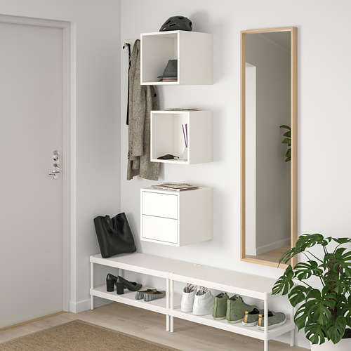Los módulos de almacenamiento EKET de pared blancos muestran y ocultan artículos. El tamaño y la forma varían, organizados junto a un espejo de cuerpo entero y una planta.