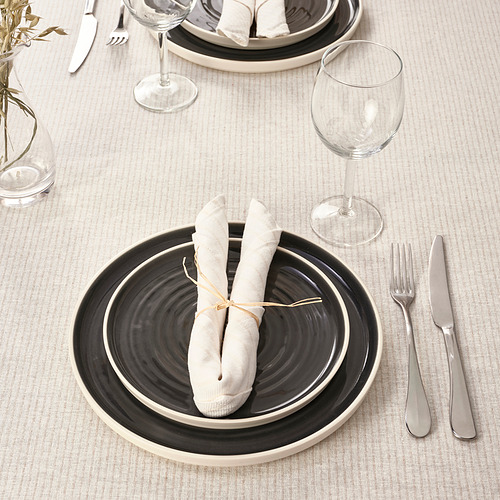 Mesa con platos blancos y negros, servilletas enrolladas, cubiertos y copas de vino sobre un mantel beige.
