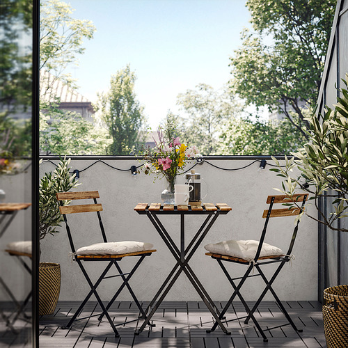 Pequeño conjunto de asientos de exterior con mesa y dos sillas, negro y de madera. Decorado con jarrón de flores y guirnaldas de luces.