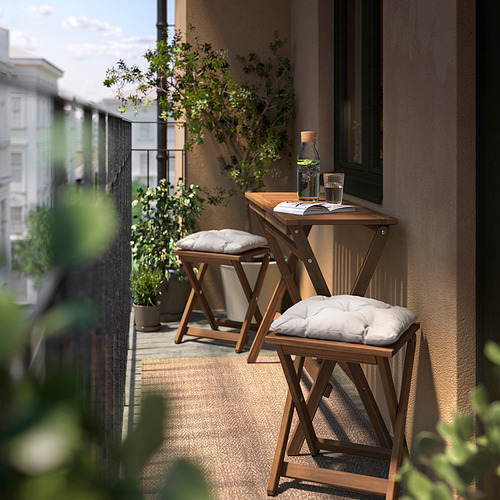 Pequeño balcón con mesa y taburetes de madera NÄMMARÖ, cojines blancos, plantas y una bebida.