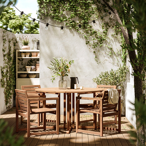 Juego de comedor al aire libre con 4 sillas y mesa de madera, bajo guirnaldas de luces y una pared blanca con vegetación.