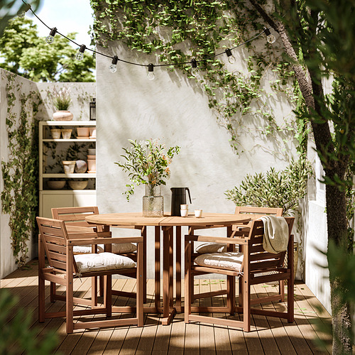 Juego de comedor al aire libre con mesa de madera y sillas acolchadas, decorado con guirnaldas de luces en lo alto, contra una pared blanca.