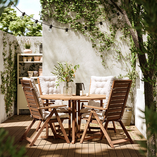 Patio exterior con mesa y sillas NÄMMARÖ, cojines. Guirnaldas de luces cuelgan en lo alto, plantas en macetas, paredes blancas.