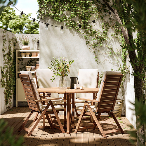 Patio al aire libre con mesa de madera, cuatro sillas a juego y guirnaldas de luces en el techo. Las sillas tienen cojines de color beige.