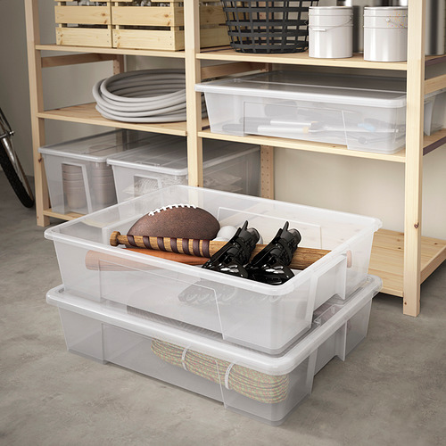 Cajas de plástico transparente SAMLA que guardan material deportivo, fútbol, patines. Caja superior más pequeña, configuración ordenada con alfombrilla a rayas. Los estantes de madera sostienen más contenedores.