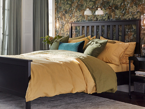 Dormitorio con cama grande, ropa de cama amarilla y verde, cabecero de madera oscura, papel pintado floral y lámparas RANARP ajustables.