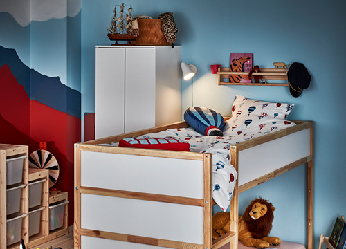 Habitación infantil con paredes azules y rojas, con una litera de madera KURA blanca con ropa de cama de colores y león de peluche.