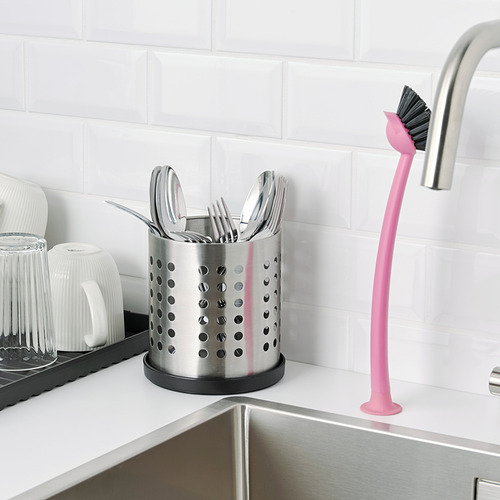 Escena de cocina con organizador de cubiertos de acero inoxidable en la encimera y cepillo rosa al lado del fregadero.