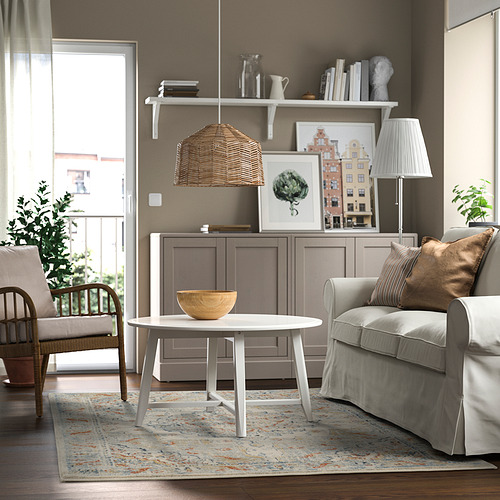 Sala de estar moderna con alfombra de colores, sillón gris, mesa de centro blanca, mesa auxiliar de madera, lámpara de mimbre y decoraciones.