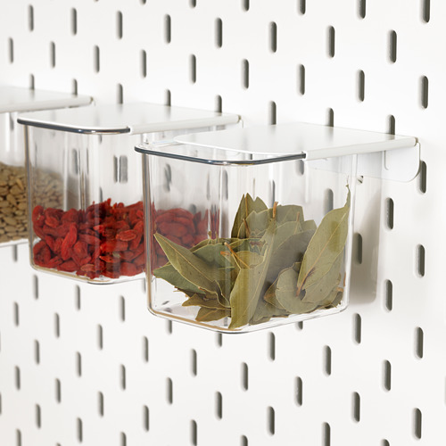 Las cajas de almacenamiento transparentes SKÅDIS cuelgan de un tablero perforado, sosteniendo especias y hojas de laurel, manteniendo los artículos organizados y accesibles.