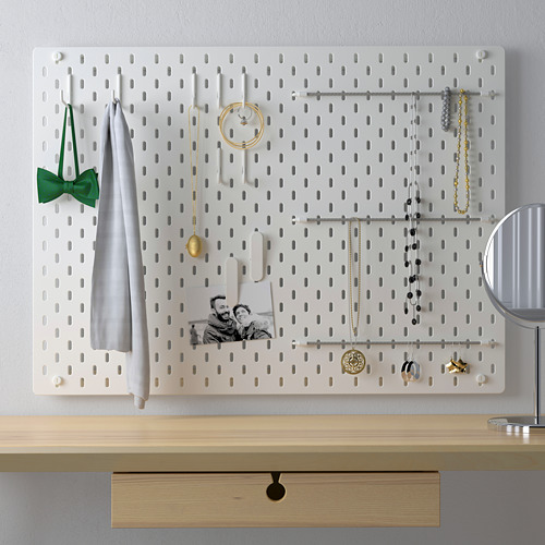Pantalla de tablero perforado blanco SKÅDIS con accesorios como joyas, telas y fotografías. Montado sobre cajón de madera, ideal para organizar objetos pequeños.