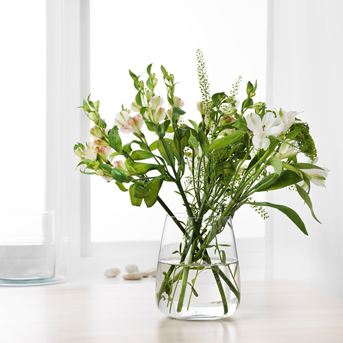 Un jarrón de vidrio transparente con flores blancas y hojas verdes sentado sobre una mesa blanca, sosteniendo agua para las flores.