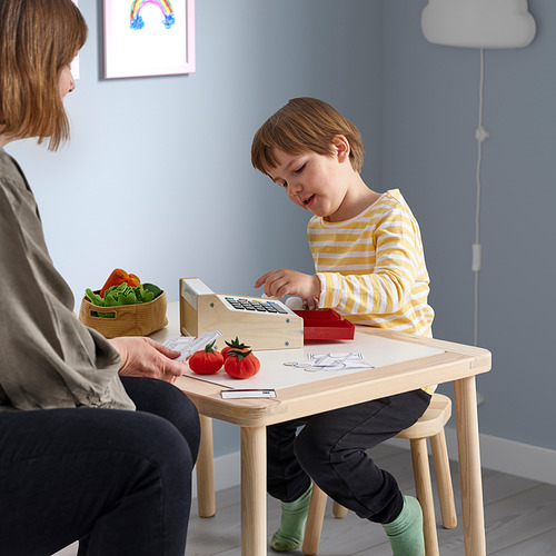 Niño jugando con la caja registradora de juguete DUKTIG en la mesa con comida de juego. Niño con camisa a rayas, con un adulto cerca.