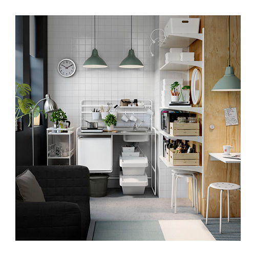 Pequeña cocina blanca con encimera gris, lámparas colgantes verdes, sofá negro y divisor de madera.