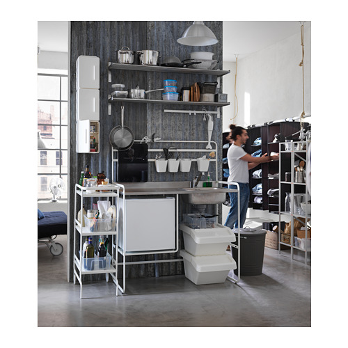 Cocina moderna y compacta con fregadero de acero inoxidable, gabinetes blancos y estanterías abiertas. Una persona está organizando objetos a su lado.
