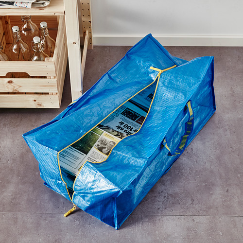 Bolsa de almacenamiento azul con asas y cremallera amarillas, tirada en el suelo gris junto a un estante de madera.