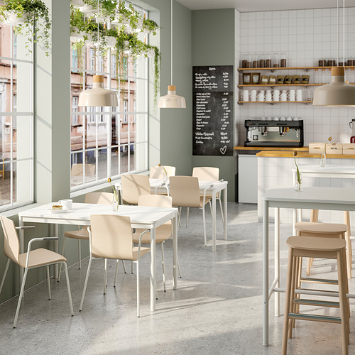 Cafetería moderna con mesas blancas y sillas de colores claros. Grandes ventanas, paredes verdes y plantas colgantes. Menú de pizarra y mostrador con cafés.