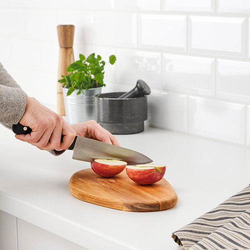 La persona corta la manzana en rodajas en la tabla de cortar de madera de acacia FASCINERA; Encimera de cocina, placa para salpicaduras de azulejos de metro blancos visibles.