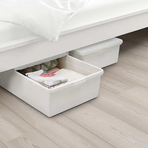Caja de plástico blanco debajo de una cama, parcialmente extraída, con ropa doblada. La tapa tiene pequeñas crestas para la circulación del aire.