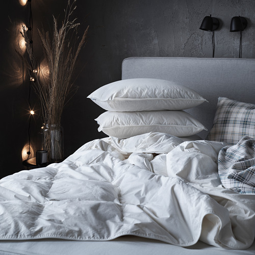 Cama deshecha con edredón blanco FJÄLLBRÄCKA; Almohadas blancas, funda de almohada a cuadros, pared oscura con plantas secas, lámparas.