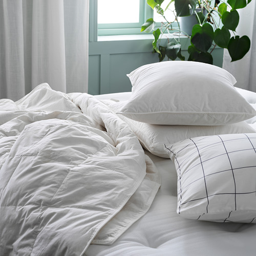 Cama blanca deshecha con edredón y almohadas, almohada con estampado de rejilla, ventana verde.