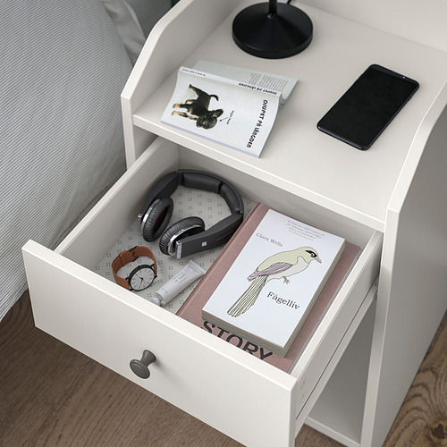 Mesita de noche blanca con estantes abiertos con libros y un teléfono inteligente, y un cajón que contiene auriculares, un pequeño reloj y un libro titulado cuento.