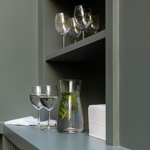 Estantería verde con copas de vino transparente, algunas llenas de agua, junto a una jarra de cristal con vegetación. A su lado se colocan servilletas blancas.