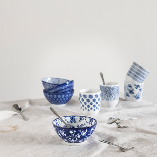 Cuencos y tazas de cerámica azul y blanca, algunos apilados, otros esparcidos sobre un mantel con cucharas.