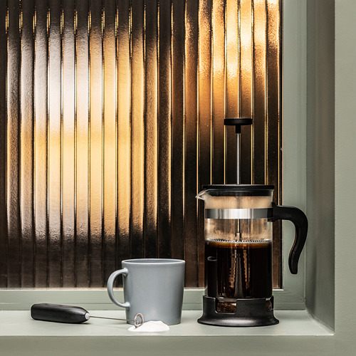 Cafetera de prensa francesa negra en el alféizar de la ventana con taza gris, varilla espumosa y bolsa de terciopelo cerca de la ventana con persianas verticales.