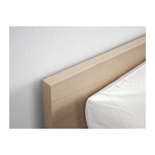 Primer plano de un cabecero de madera clara contra una pared blanca, de líneas limpias y acabado liso, con una esquina de una almohada blanca a la vista.