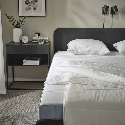 Escena de dormitorio con edredón SANDGRÄSMAL en la cama, mesita de noche gris con libros, reloj y planta; Cabecero negro, apliques.
