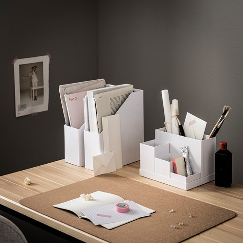 Ordenada configuración de escritorio con organizadores blancos, libros, papeles y una almohadilla de escritorio de corcho, contra una pared oscura con una botella negra y una imagen de arte.