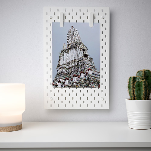 El tablero perforado SKÅDIS blanco muestra una foto grande, rodeada de agujeros espaciados uniformemente. Montado en una pared de luz, con una planta de cactus y una lámpara cerca.