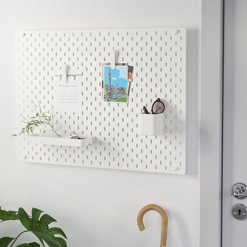 El tablero perforado SKÅDIS blanco con ganchos, taza, planta, calendario y foto sostiene varios artículos de manera ordenada. Ideal para organizar entradas u oficinas.