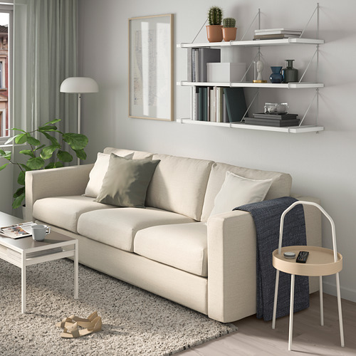 Salón moderno con sofá BERGSHULT color crema, estantería de pared con libros y plantas, mesa auxiliar con mando a distancia y lámpara.