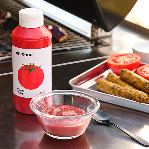 Descripción de la imagen: una botella roja de KETCHUP junto a un recipiente de vidrio con KETCHUP, salchichas y tomates en rodajas en una bandeja, con una cuchara.