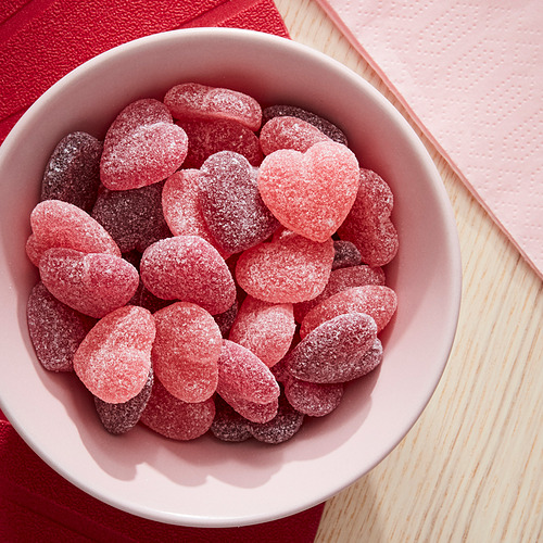 Cuenco blanco con caramelos de corazón rosas recubiertos de azúcar. Los caramelos son rojos y rosas, esparcidos en el tazón. Cuenco sobre una superficie de madera con paño rojo.