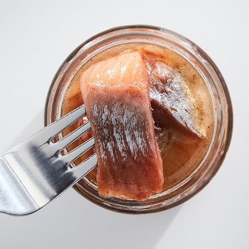 Un frasco de arenque marinado de color rosa claro, sostenido por un tenedor. Recipiente de vidrio transparente, tenedor de plata.